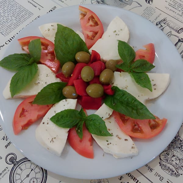 Caprese salad with mozzarella, tomatoes, basil, olives, and red peppers on a white plate.