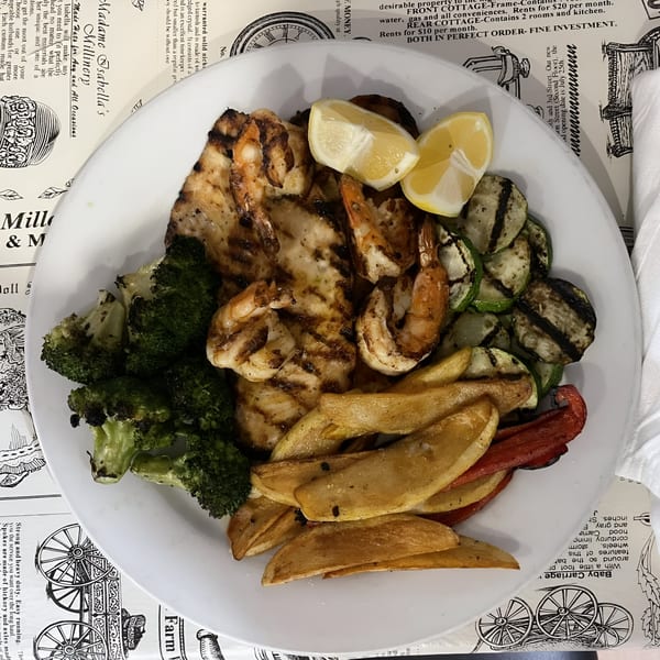 Grilled chicken and shrimp with lemon wedges, broccoli, zucchini, and potato wedges on a white plate.