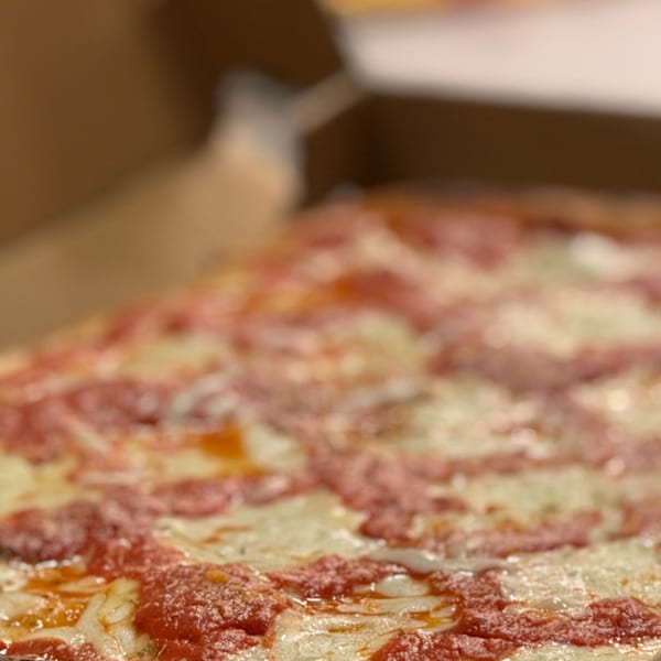 Close-up of a square-cut Angelo's Pizzeria pizza with melted cheese and red sauce in a box.