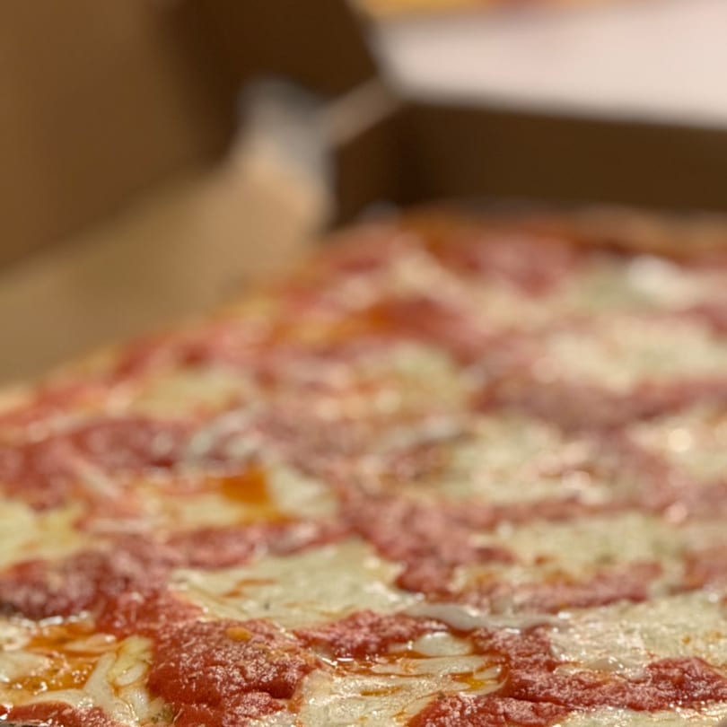Close-up of a square-cut Angelo's Pizzeria pizza with melted cheese and red sauce in a box.